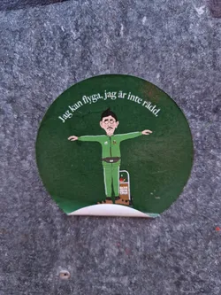 Street sticker A round green sticker with an illustration of a man with his arms outstretched, as if flying. The text around the illustration says: "Jag kan flyga, jag är inte rädd" which translates to  "I can fly, I'm not afraid" in Swedish. The man is wearing green clothes and a belt bag.