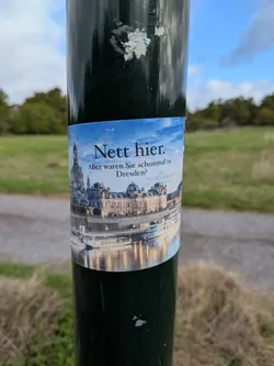 Street sticker A rectangular sticker affixed to a dark green metal pole. The sticker features a scenic photograph of the Dresden skyline along the Elbe river, showcasing historic architecture including the Frauenkirche dome. The sky is blue with soft white clouds. Overlaid on the sky is black text in a serif font that reads 'Nett hier. Aber waren Sie schonmal in Dresden?' in German, mimicking the famous Baden-Württemberg tourism slogan.