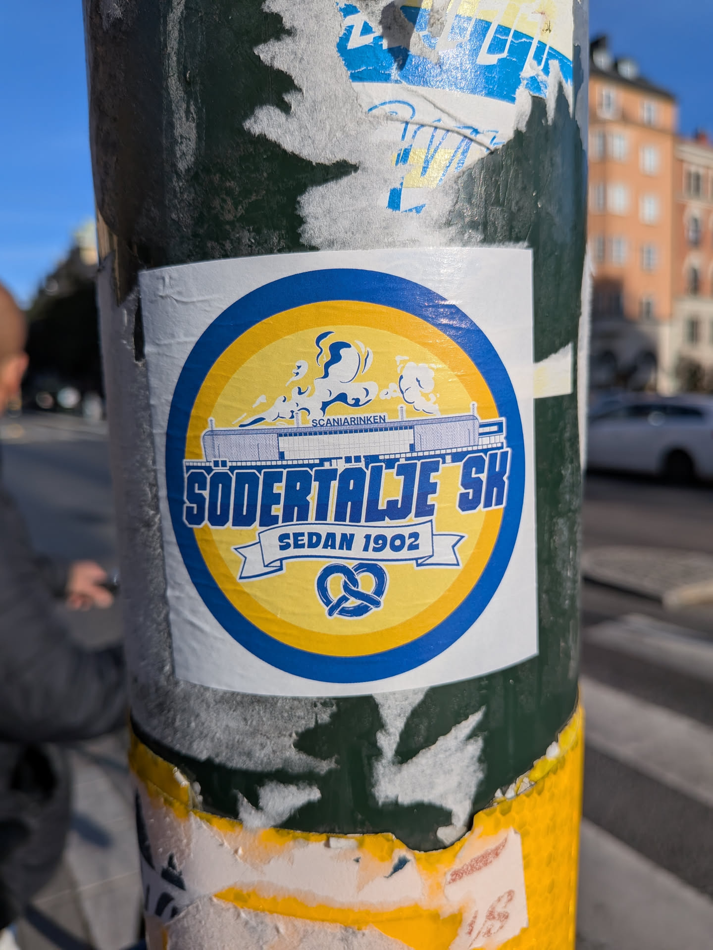 The sticker is circular with a blue and yellow color scheme. The outer ring is blue, followed by a yellow inner ring. In the center, there is a depiction of a building or structure with the text 'SCANIARINKEN' above it. Below this, in large blue letters, is 'SÖDERTÄLJE SK'. A white banner with the text 'SEDAN 1902' is positioned below the team name. At the bottom of the sticker, there is a blue pretzel symbol. The sticker appears to be slightly worn and is affixed to a pole.