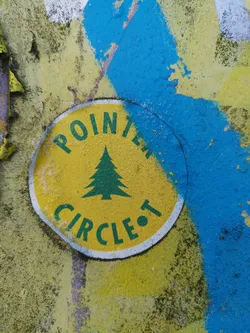 A round yellow sticker with a simple design.  It features the words 'POINT' and 'CIRCLE' in green text, with a small green evergreen tree image in between. The sticker appears to be affixed to a textured surface with blue and yellow paint. 