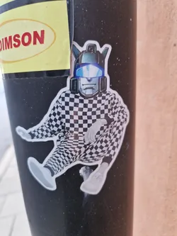 Street sticker A sticker featuring a stylized Transformer character in a checkered jumpsuit. The character's head is clearly that of a Transformer, while the body is dressed in a black and white checkered outfit. The character is depicted in a relaxed, almost lounging pose.