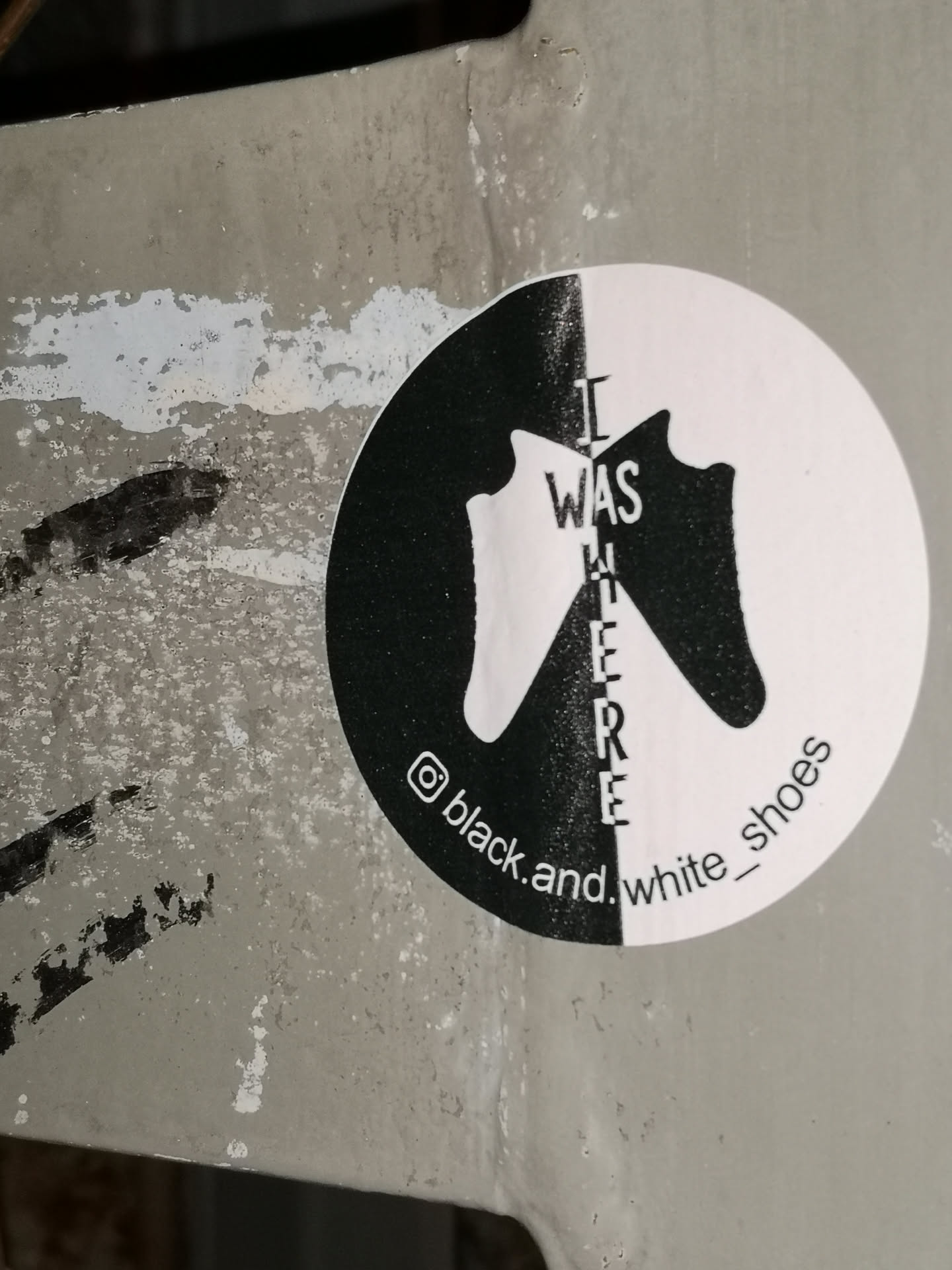 A circular sticker featuring a minimalist design split vertically into black and white halves. On the left, a white shoe silhouette is set against the black background, while on the right, a black shoe silhouette is set against the white background. The two shoes face each other, creating a central negative space. Vertically aligned in the center, the text 'I WAS HERE' is displayed, with each letter split by the black and white division. Along the bottom curve, an Instagram logo precedes the handle '@black.and.white_shoes'. The sticker has a slightly textured appearance, suggesting it's adhered to a weathered surface.