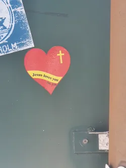 A red heart-shaped sticker with a yellow banner at the bottom and a yellow cross at the top. The text on the sticker says "Jesus loves you" and "John 3:16".