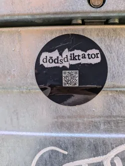A black round sticker is affixed to a metallic surface. The sticker has a white text "dödSdiktator" and a QR code below it. The QR code is scannable.