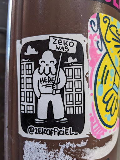 Street sticker Black and white sticker with a cartoon character holding a sign that says "Zeke was here." The character is standing in front of buildings. The sticker also includes the artist's Instagram handle.