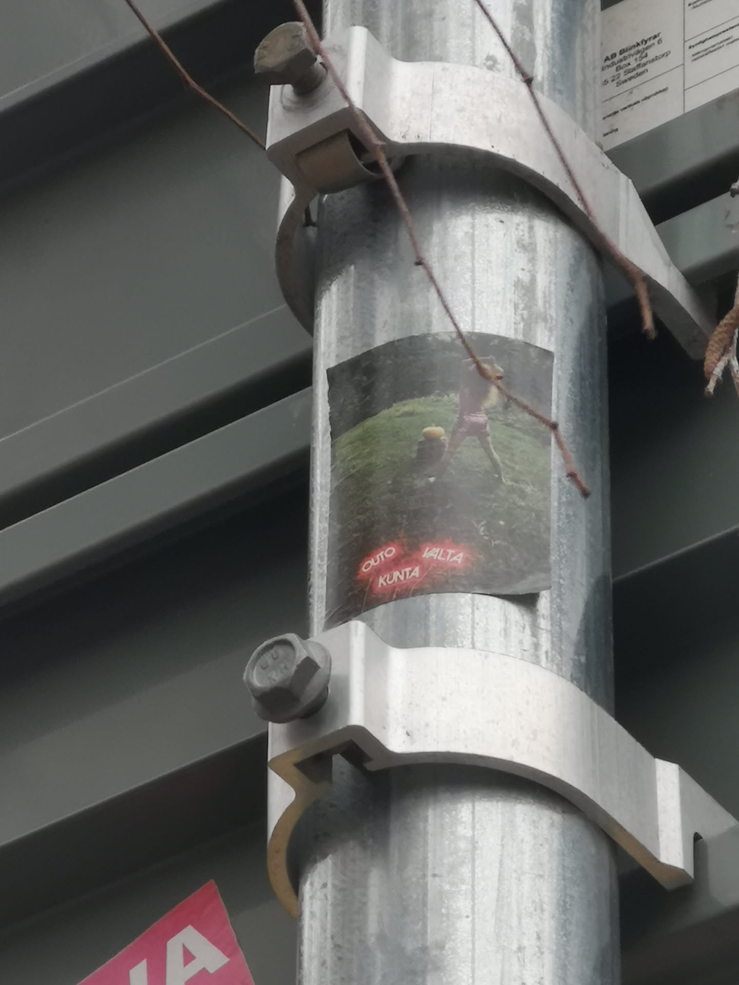The sticker is a rectangular print adhered to a galvanized metal pole. It features a dark, somewhat blurry image of a person, possibly wearing light pink shorts and a top, standing in a green, grassy landscape. The person appears to be in motion, perhaps swinging an object. Near the bottom left, three words are prominently displayed in a glowing red, neon-like font: "OUTO", "VALTA", and "KUNTA". The overall aesthetic is grainy and slightly mysterious, resembling a lo-fi photograph or a meme.