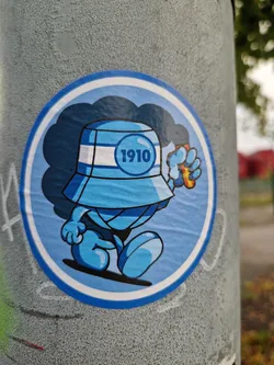 Street sticker A round sticker featuring a cartoon character wearing a blue bucket hat with the number 1910 on it. The character is light blue and appears to be walking. It is holding a small, orange flame-like object in its hand. The background is a dark blue cloud-like shape.