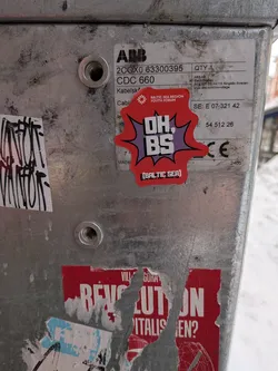 Street sticker The sticker is an irregularly shaped, vibrant red starburst or speech bubble design, outlined in black. It features a purple inner burst, against which the main text 'OH. BS' is prominently displayed in large, bold white letters with a black outline, in a comic book style font. Above this, in smaller white text, is 'BALTIC SEA REGION YOUTH FORUM' accompanied by a small, stylized logo resembling a snowflake or star. Below the main text, in white bracketed text, is '[BALTIC SEA]'. The sticker has a dynamic and impactful visual style.