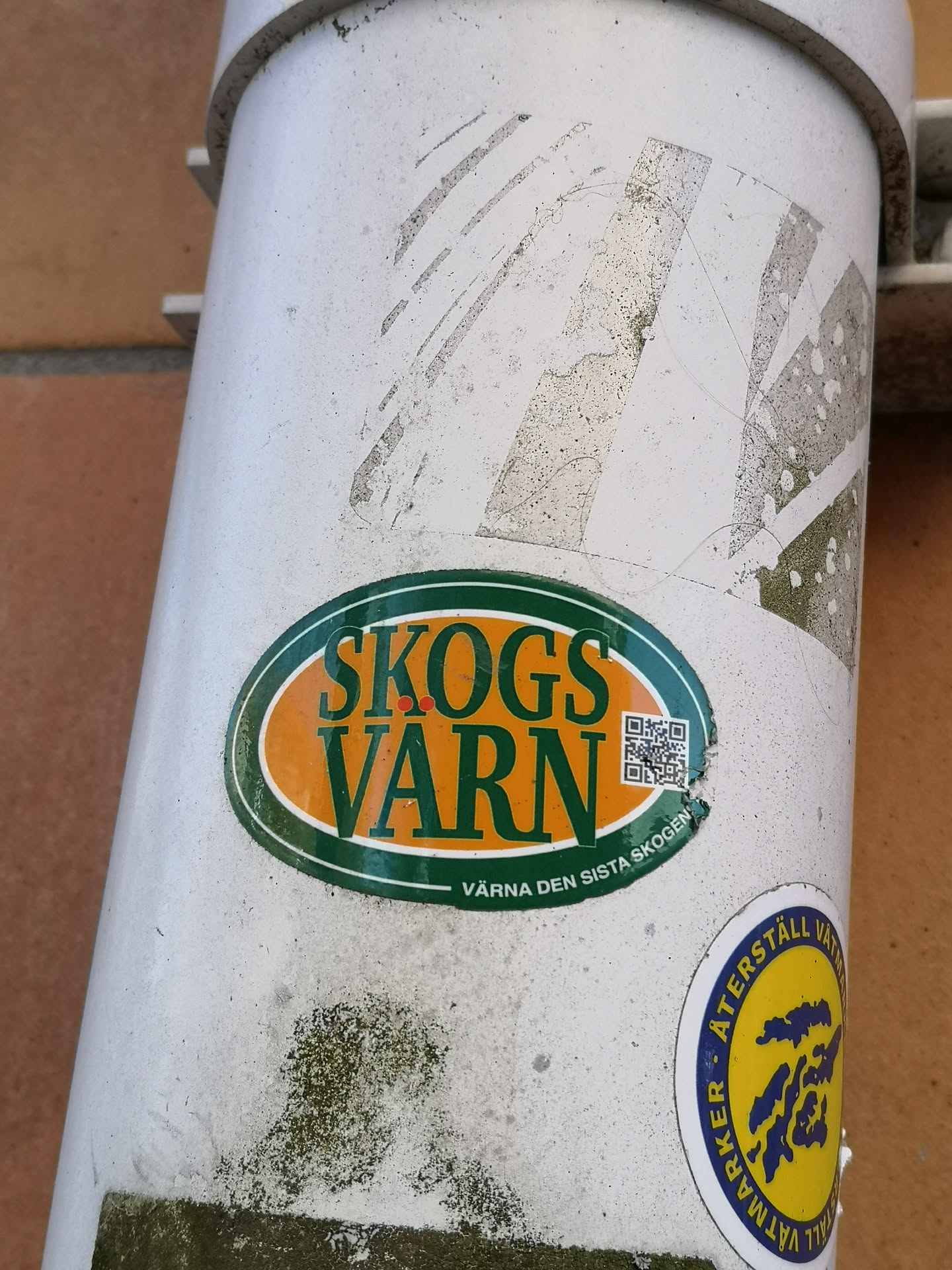 An oval-shaped sticker with a dark green border, featuring a vibrant orange background. The main text "SKOGS VARN" is prominently displayed in bold, dark green letters. Below this, in smaller dark green text, reads "VÄRNA DEN SISTA SKOGEN". To the right of the main text, within the orange area, there is a small, black and white QR code. The sticker shows signs of wear and tear, with some peeling and dirt accumulation, indicating its age and exposure to elements.