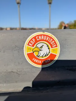 Street sticker A round sticker with a yellow and orange border. The central image features a yellow and white eagle head inside a yellow circle.  The text 'KOP CHAUVINOIS' is printed in a semicircle above the eagle, and 'FORZA L'USC' is printed below. The numbers '20' and '21' are to the sides of the eagle.
