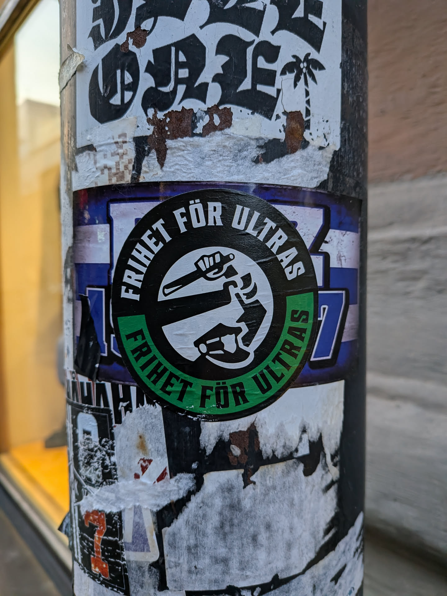 The sticker is circular with a black border. The outer ring of the circle is green and contains the text 'FRIHET FÖR ULTRAS' in white capital letters. The inner part of the circle is white and features a black graphic. This graphic depicts a clenched fist holding a baton or club, raised upwards. Below the fist, there is a stylized representation of a car or vehicle. The sticker is placed on a pole that is covered in many other torn and weathered stickers, with some blue and white striped background visible behind the central sticker.