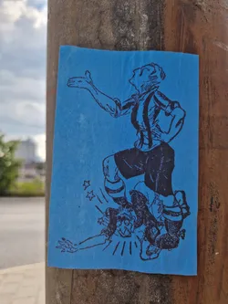 Street sticker A blue rectangular sticker featuring a black and white drawing of a soccer player performing a celebratory move by stepping on their fallen opponent. The sticker has a slightly worn look, suggesting it may have been affixed outdoors for some time. The style is reminiscent of vintage sports cartoons or illustrations.