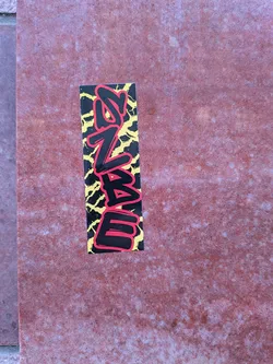 The sticker is rectangular and oriented vertically. It features stylized, bold lettering in black with a red outline. The background is a chaotic pattern of jagged yellow lines on a black base, resembling lightning or cracks. The overall aesthetic is energetic and urban.