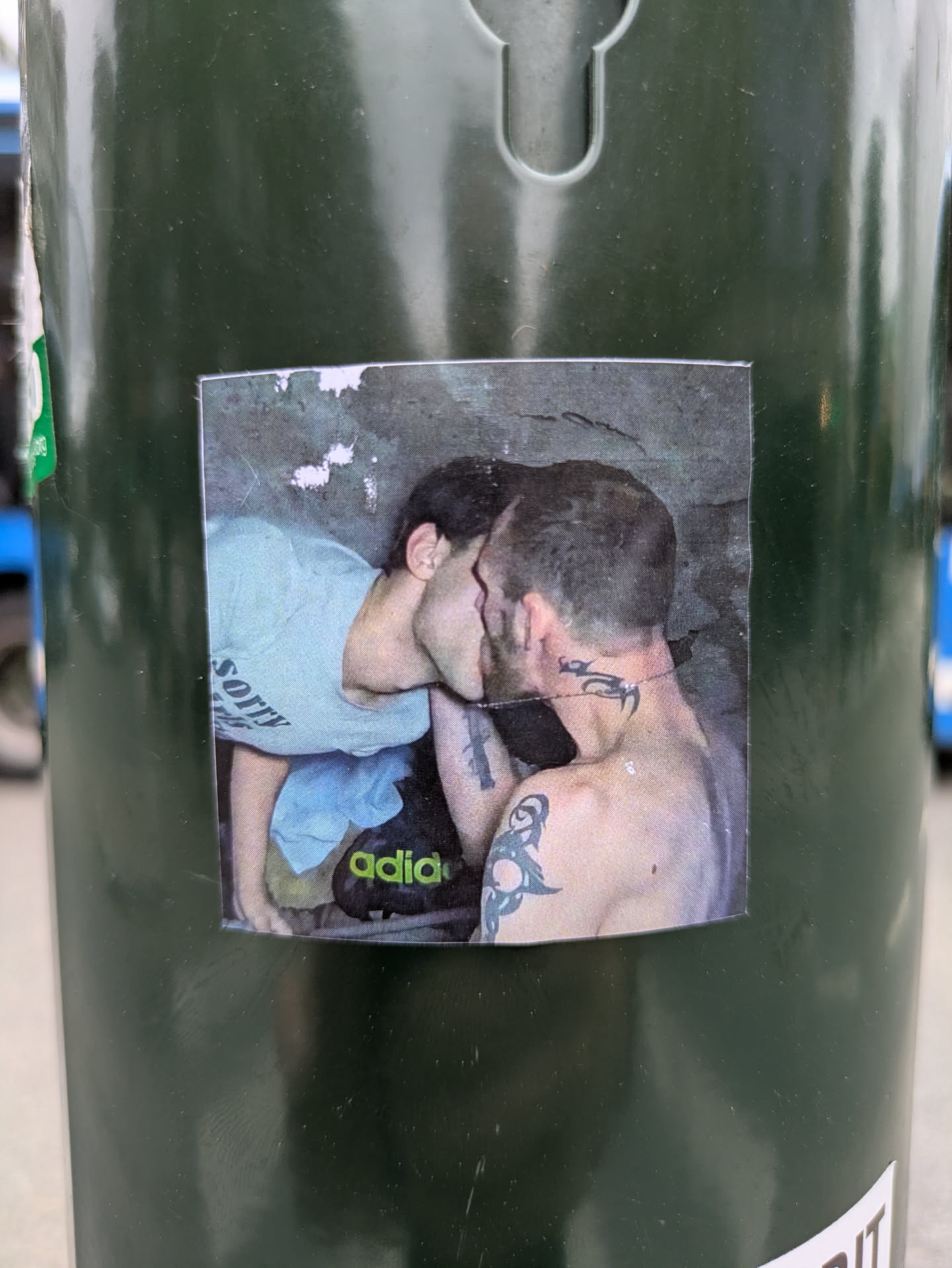 A rectangular, printed sticker affixed to a dark green pole or bin. The sticker features a photograph of two individuals, seemingly male, in a close embrace and kissing. The person on the left is wearing a light-colored t-shirt with the word 'SORRY' visible on the back. The person on the right has visible tattoos on their left arm and neck/shoulder area, which appear to be tribal or abstract designs. A green 'adidas' logo is visible on what looks like a bag or shoe in the lower part of the image. The background is a textured, possibly concrete or rock surface, with some lighter patches.