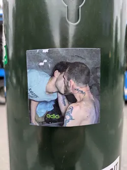Street sticker A rectangular, printed sticker affixed to a dark green pole or bin. The sticker features a photograph of two individuals, seemingly male, in a close embrace and kissing. The person on the left is wearing a light-colored t-shirt with the word 'SORRY' visible on the back. The person on the right has visible tattoos on their left arm and neck/shoulder area, which appear to be tribal or abstract designs. A green 'adidas' logo is visible on what looks like a bag or shoe in the lower part of the image. The background is a textured, possibly concrete or rock surface, with some lighter patches.
