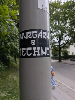 Street sticker A black rectangular sticker with bold white text reading 'DJURGÅRN & TECHNO' adhered to a grey metal pole outdoors.