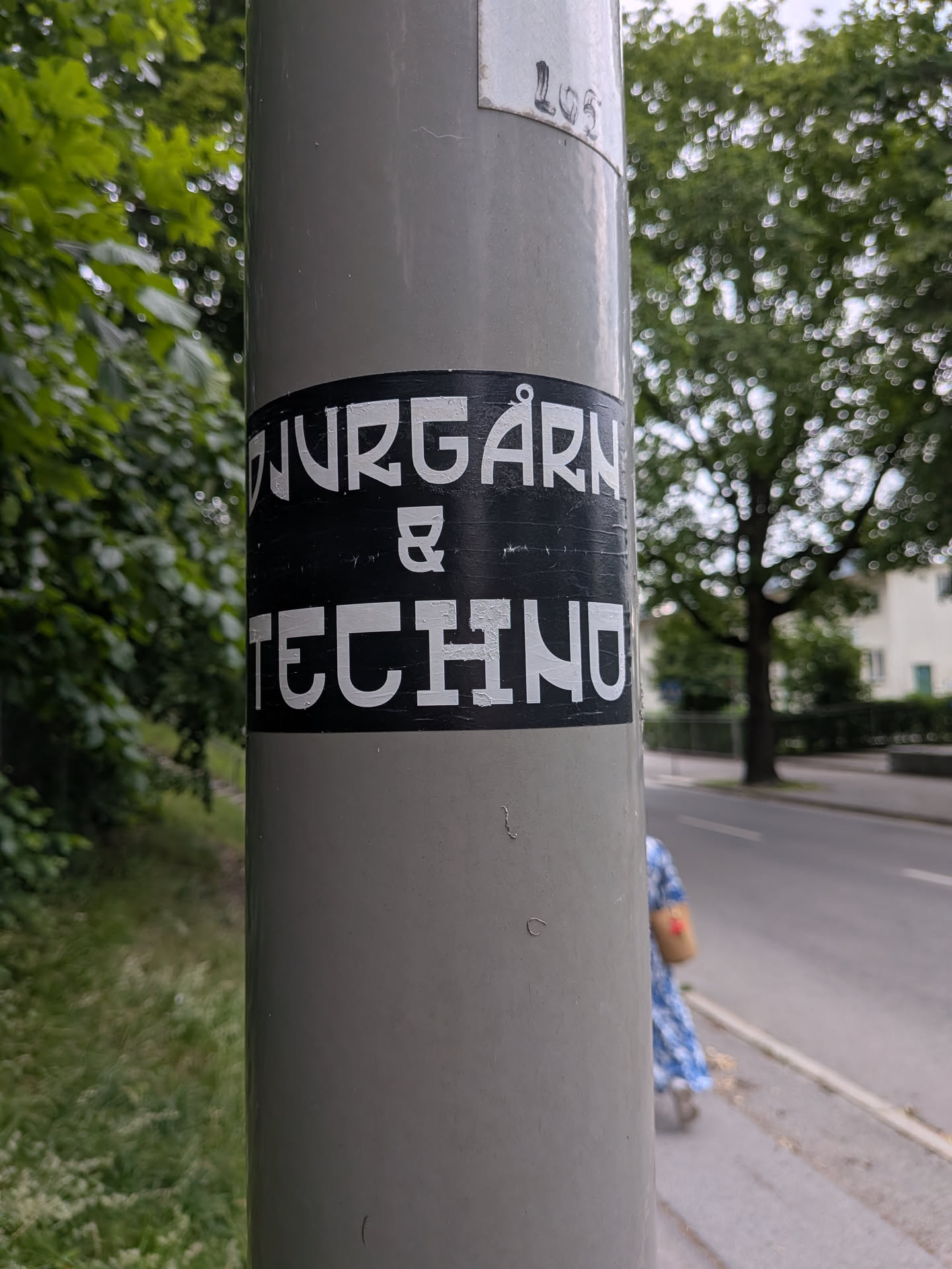 A black rectangular sticker with bold white text reading 'DJURGÅRN & TECHNO' adhered to a grey metal pole outdoors.