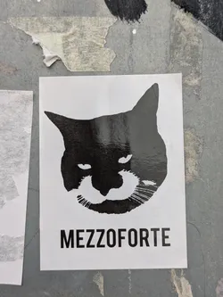 Street sticker This black and white sticker displays a stylized image of a cat's head or a similar creature with feline features. The design is simple yet striking, and the text "MEZZOFORTE" is printed in bold, sans-serif font underneath the illustration. The sticker appears to be placed outdoors, adhered to a weathered or textured surface.