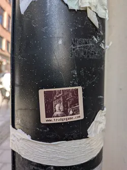Street sticker A rectangular sticker with rounded corners on a cream-colored background. It features a dark brown, detailed illustration in a woodcut or pixel-art style, depicting a hooded figure from behind looking at a complex control panel or machinery. Below the illustration, the website address 'www.trudgegame.com' is printed in a dark brown, lowercase font.