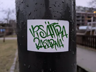 Street sticker A close-up shot of a white sticker on a dark pole. The sticker features green graffiti-style lettering that appears to say "VISA HIRA RASARMA". The pole is wet, with water droplets visible on its surface. The background is slightly blurred but shows some outdoor scenery, such as trees and buildings in the distance.
