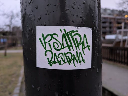 Street sticker A close-up shot of a white sticker on a dark pole. The sticker features green graffiti-style lettering that appears to say "VISA HIRA RASARMA". The pole is wet, with water droplets visible on its surface. The background is slightly blurred but shows some outdoor scenery, such as trees and buildings in the distance.