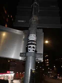 Street sticker The sticker is vertically oriented, predominantly black and white, and wrapped around a metal pole. The top section features a cartoonish, wide-eyed face with a distressed or surprised expression, mouth agape, rendered in white on a black background. Below this, a black band contains white capital letters spelling 'SUPPORT GR-VIT', with the 'A' and 'Y' of 'GRAVITY' seemingly cut off or obscured. The bottom section displays a detailed, realistic-looking skull, also in white on a black background, showing intricate bone structure, eye sockets, and teeth.