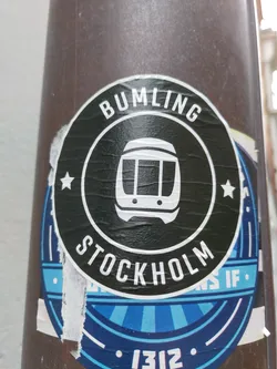 A black and white round sticker with the text "BUMLING" and "STOCKHOLM" in a circular pattern. There is an icon of a train in the center. The sticker is attached to a dark brown pole.