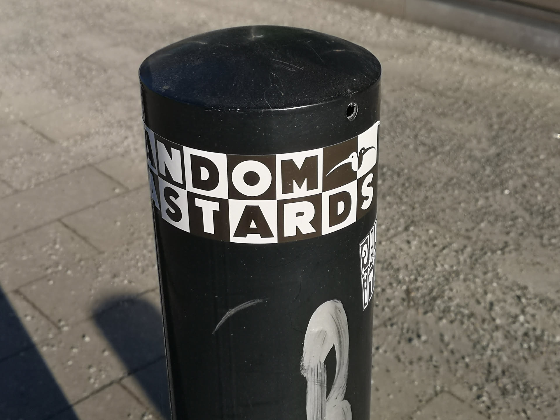 The sticker is a rectangular band wrapped around a black cylindrical bollard. It features bold, uppercase, sans-serif text 'RANDOM BASTARDS' in white. The background behind the letters is a checkerboard pattern of alternating black and white squares. A stylized white silhouette of a kiwi bird is integrated into the design, replacing part of the 'M' in 'RANDOM' and extending over the 'S' in 'BASTARDS'. The overall aesthetic is stark and graphic, primarily in black and white.