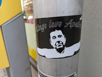 Street sticker This sticker shows a stylized black and white portrait of a man's face with a surprised or slightly fearful expression, set against a black background.  Above the portrait is the phrase "Linge leve Apetos" written in a cursive script.