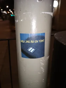 Street sticker The sticker is rectangular with a dark blue background. It features a close-up image of a dark blue, possibly black, knitted beanie. On the beanie, there is a distinct logo: a diamond shape with two horizontal white stripes against a blue background. Above the beanie image, in the upper left portion of the sticker, yellow text is prominently displayed. The sticker itself shows signs of wear, with slight peeling at the edges and minor surface scratches, indicating it's a physical sticker adhered to a light-colored pole.