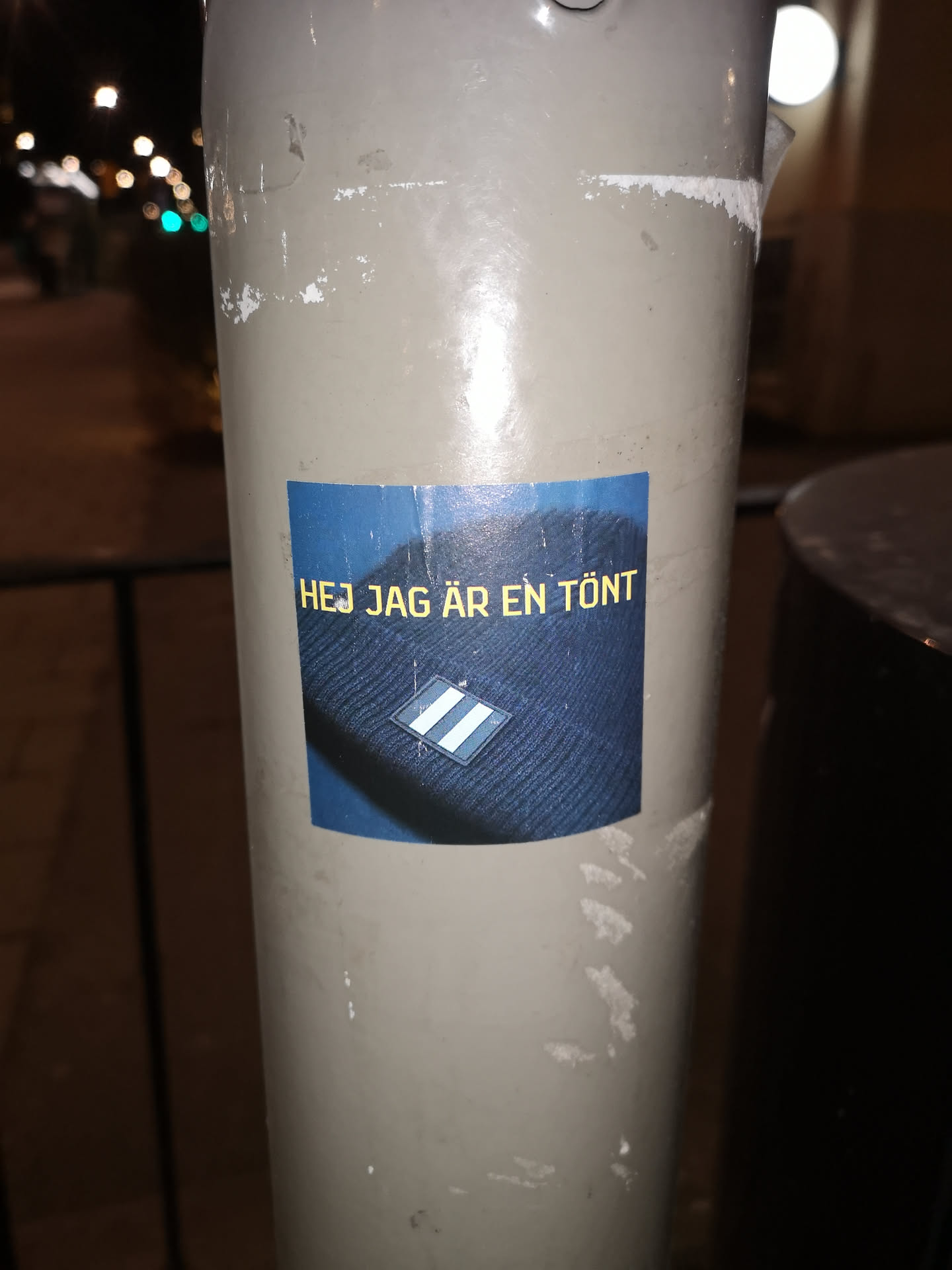 The sticker is rectangular with a dark blue background. It features a close-up image of a dark blue, possibly black, knitted beanie. On the beanie, there is a distinct logo: a diamond shape with two horizontal white stripes against a blue background. Above the beanie image, in the upper left portion of the sticker, yellow text is prominently displayed. The sticker itself shows signs of wear, with slight peeling at the edges and minor surface scratches, indicating it's a physical sticker adhered to a light-colored pole.