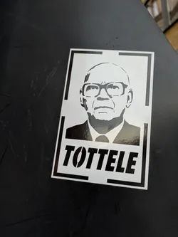 Street sticker The sticker features a black and white stencil portrait of a bald man wearing glasses and a suit. The portrait is rendered with simple, bold lines, and the text 'TOTTELLE' is prominently displayed in stenciled capital letters below the image. The sticker has a white background with a thin black border, and a thicker black border framing the entire design.