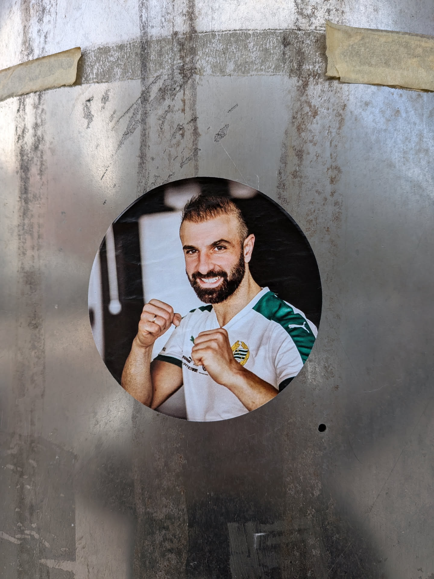 The sticker is circular, depicting a smiling man with a dark beard and short, styled hair. He is shown from the chest up, wearing a white sports jersey with green accents on the shoulders and sleeves. His hands are raised in a boxing or fighting stance, with fists lightly clenched. A gold and black crest-like logo is visible on the left side of his chest, and text like 'PUMA' and 'INTERSPORT' can be partially seen on his shirt. The background behind him is dark and slightly blurred, suggesting an indoor setting like a gym.