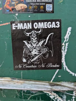 Street sticker A square black sticker with white graphics and text, pasted on a green surface. At the top, the words 'E-MAN OMEGA3' are printed in a bold, white, sans-serif typeface. The central illustration depicts a skeletal warrior in detailed armor, wearing a horned helmet and holding a large battle axe. The armor features skull designs on the shoulder plates. At the bottom of the sticker, the phrase 'No Countries No Borders' is written in an elegant, white cursive script.