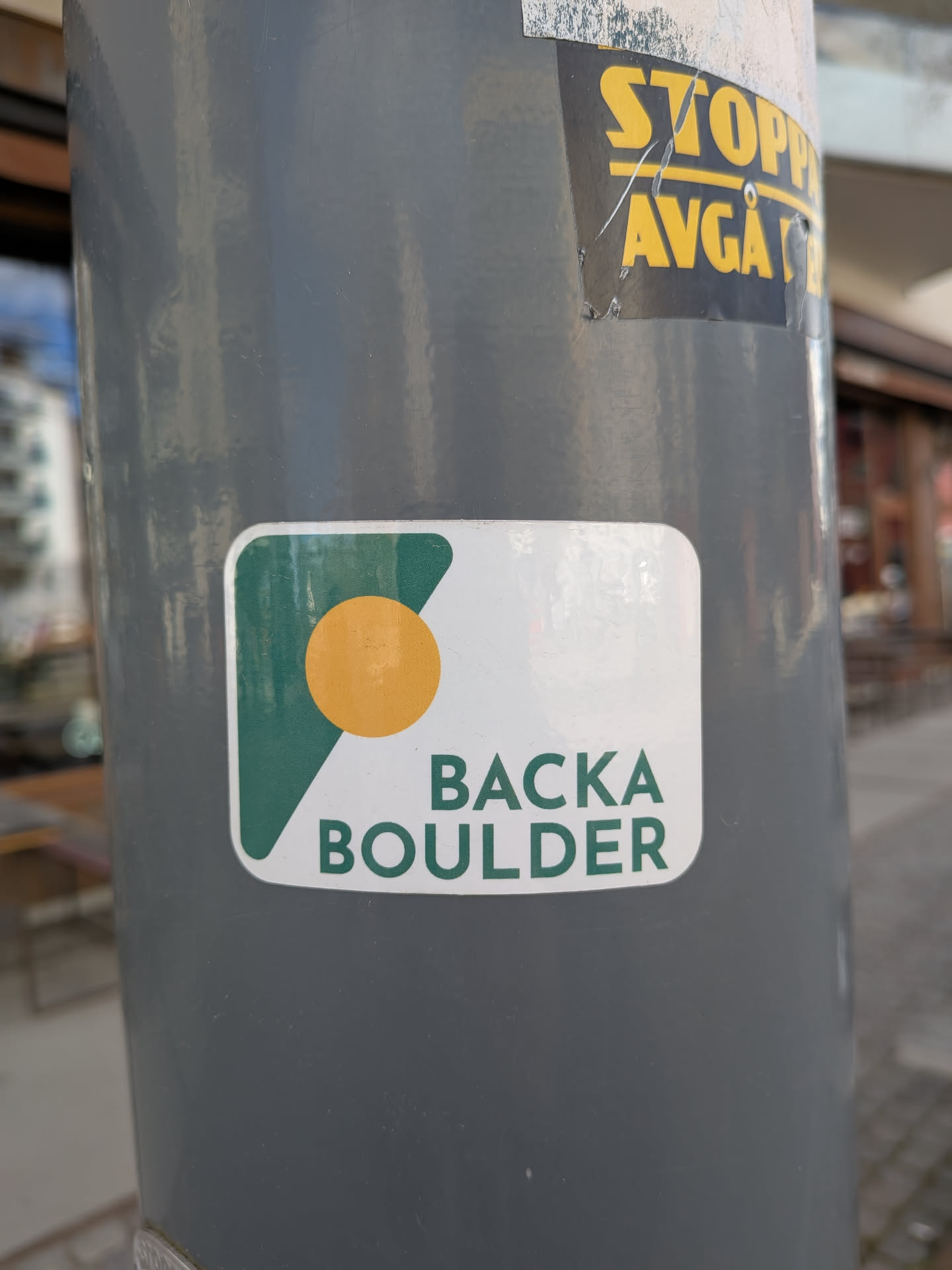 A rectangular sticker with rounded corners, featuring a clean, modern design. The background is white. On the left side, there's a dark green, angular shape that resembles a stylized 'B' or a rock formation, with a solid orange circle placed within its upper left portion. To the right of this graphic, the text 'BACKA' is stacked above 'BOULDER' in a bold, sans-serif font, both in the same dark green color as the geometric shape.
