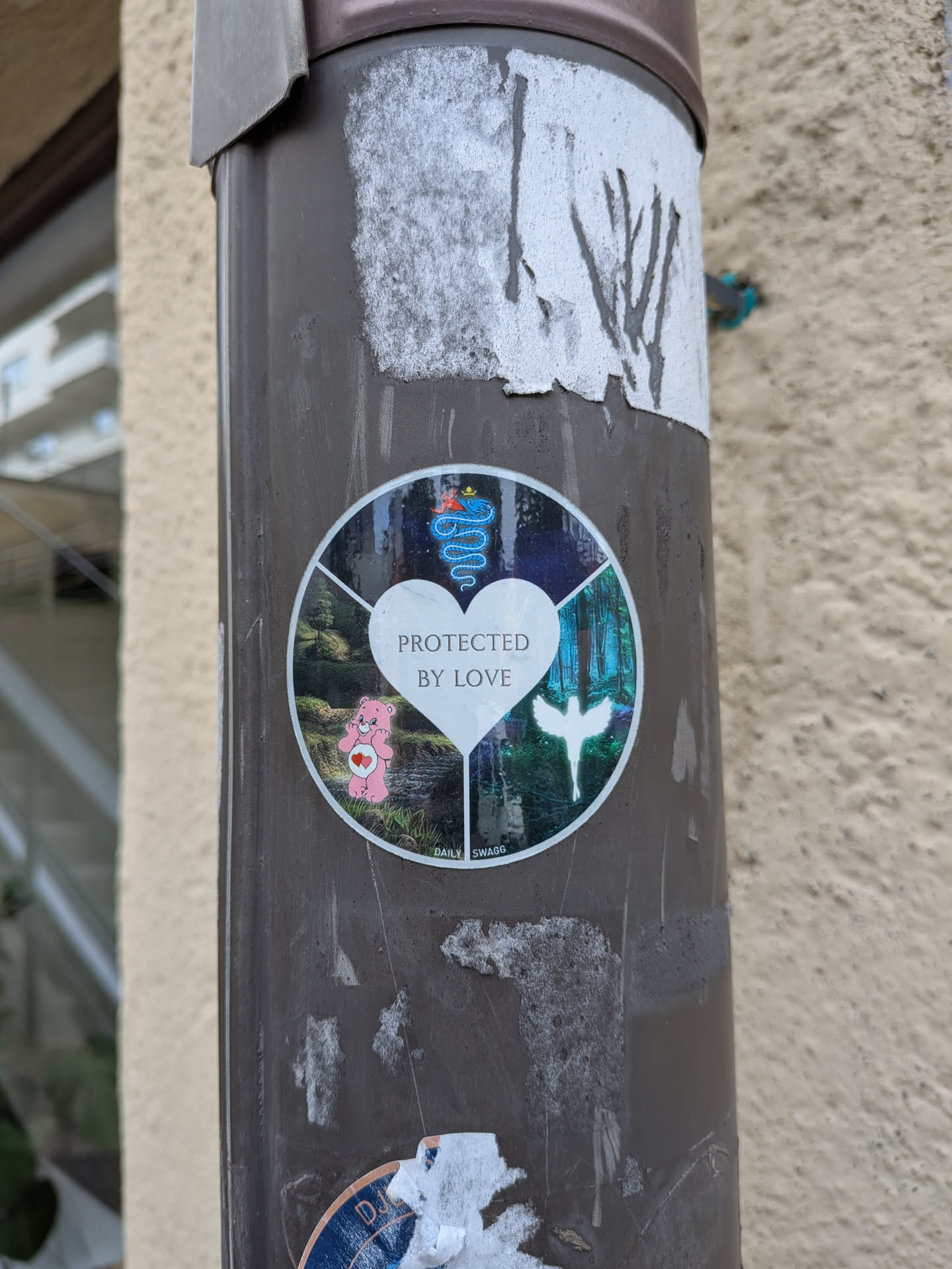 A round sticker with a central white heart containing the text 'PROTECTED BY LOVE'. The background is split into four fantasy-themed segments featuring a pink Care Bear, a crowned blue snake, a glowing angel, and a dark forest scene.