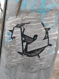 A partially damaged sticker with an abstract design in black and white. The sticker appears to be affixed to a weathered surface with other markings visible in the background. The central design is reminiscent of a stylized figure or letter.