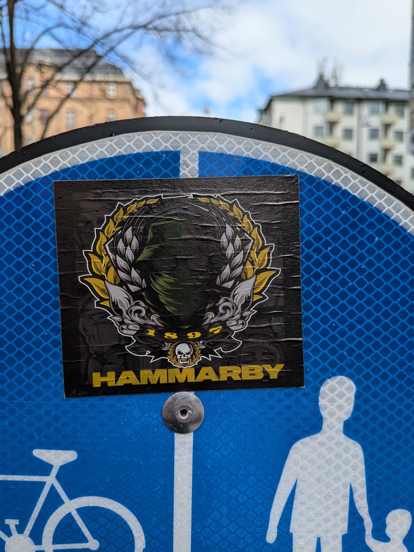 A rectangular sticker with a predominantly black background, featuring a central emblem. The emblem depicts a masked figure, possibly wearing a green balaclava or scarf, framed by a split laurel wreath. One half of the wreath is golden-yellow, and the other is silver-grey. Below the masked figure, within the wreath, is a small skull emblem, also adorned with a miniature golden wreath, and the year '1897'. At the bottom of the sticker, in bold yellow capital letters, is the word 'HAMMARBY'. The sticker appears slightly wrinkled and worn, indicating it's a physical object adhered to a surface.