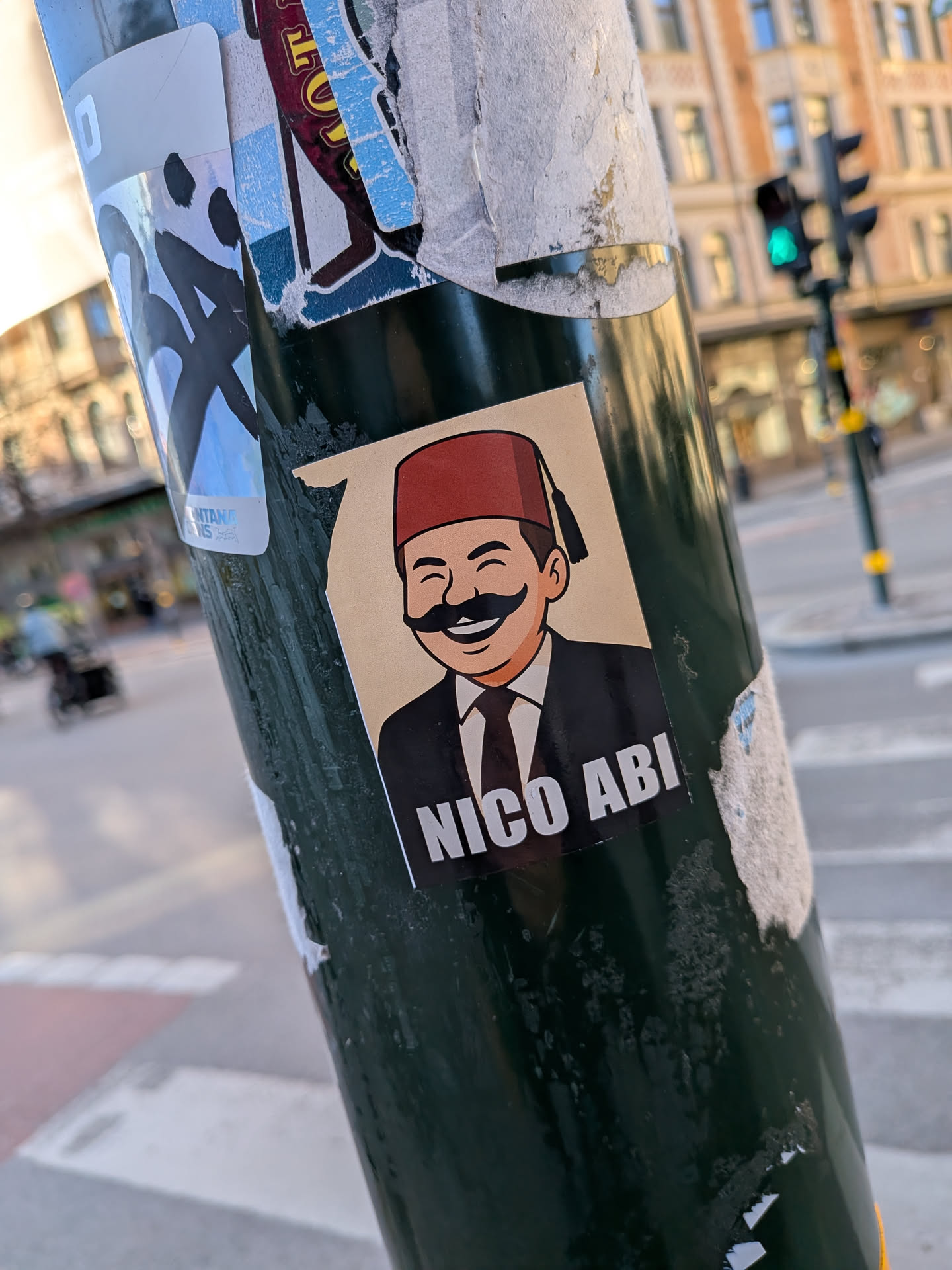 The sticker features a cartoon illustration of a smiling man with a light skin tone and a prominent, thick black mustache. He is depicted wearing a red fez with a black tassel on his head, and a black suit jacket over a white shirt with a dark tie. The background of the illustration is a plain light beige or tan color. Below the character, there is a black rectangular bar with the text "NICO ABI" written in white, bold, capital letters. The sticker has a clean, graphic art style.