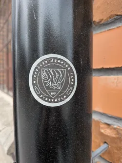 Street sticker The sticker is circular with a white border and a white outline. The center features a shield-like emblem with a stylized depiction of running legs in white lines. Above the emblem, the text 'MIKKELER RUNNING CLUB' is written in a circular fashion. Below the emblem, the year '2017' is prominently displayed, and further down, 'STOCKHOLM' is also written in a circular manner. Small icons resembling beer bottles are placed on either side of the emblem within the circular text.