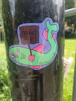 Street sticker A charming sticker featuring a cartoon fish holding a sign that says "Howet dör!" in Swedish. The fish has a light green body, pink fins, and a sign in its mouth.  The artist's signature, Filip Rahim Hansson, is visible at the bottom.