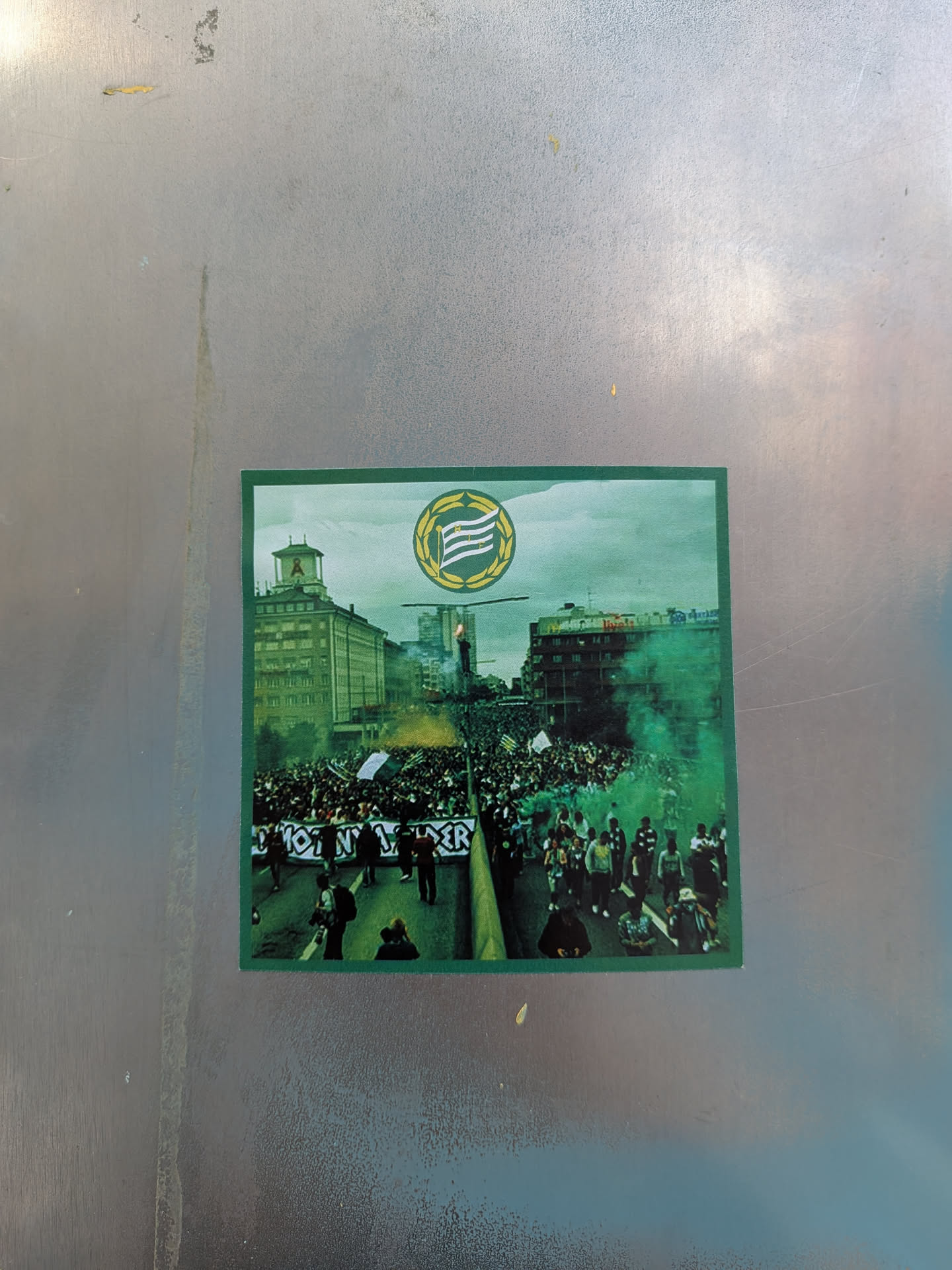 The sticker depicts a dynamic scene of a large crowd gathered in a city street or square, viewed from a slightly elevated perspective. Dominant green smoke billows from the center of the crowd, creating a dramatic atmosphere. In the background, several multi-story buildings are visible, one with a distinctive tower. At the top center of the sticker, a circular emblem features a yellow wreath encircling horizontal green and white stripes. A banner held by individuals in the foreground is partially visible. The overall color palette is dominated by greens, greys, and the muted tones of the urban environment.