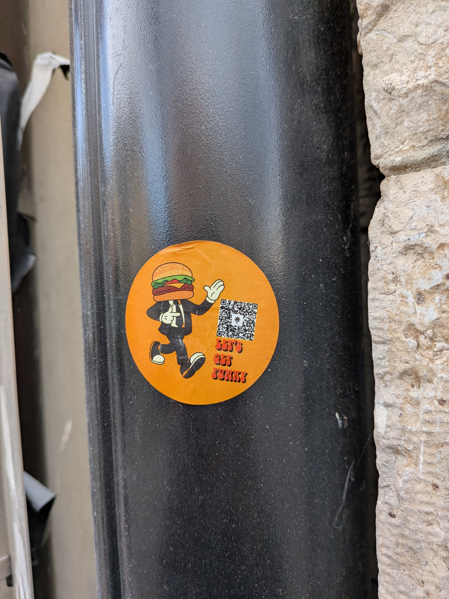 A vibrant, circular orange sticker featuring a cartoon character with a hamburger for a head, wearing a black suit, white shirt, and black tie. The character is depicted in a dynamic, running or dancing pose with white gloves and black shoes. The hamburger head includes a bun with sesame seeds, a patty, lettuce, and tomato. To the right of the character, there is a black and white QR code with a small white asterisk-like symbol in its center. Below the QR code, the text 'LET'S GET FUNKY' is written in a stylized, orange font with a darker outline. The overall design is playful and energetic.