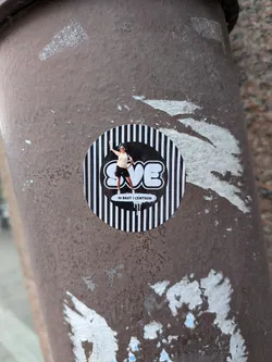 Street sticker A round sticker with a black and white vertically striped background. A central black circle features the white bubble-style text 'SVE' with a cutout photo of a person waving. Below, a white paint-drip graphic contains the text '50 BAST I CENTRUM'.