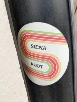 Street sticker A round sticker with a retro-style design. The sticker features multiple colored stripes, with the colors in the order: green, orange, red, orange forming an S shape. The text "SIENA" and "ROOT" are written in black in between the stripes.