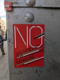 Street sticker The sticker is predominantly red with white lettering. The word 'NO' is prominently displayed in large, stylized white block letters. Below 'NO', the text 'JUSTIFICATION FOR GENOCIDE' is written in smaller, black, stencil-like block letters. Diagonal lines in green, white, and black are layered across the lower portion of the sticker, adding a graphic element.