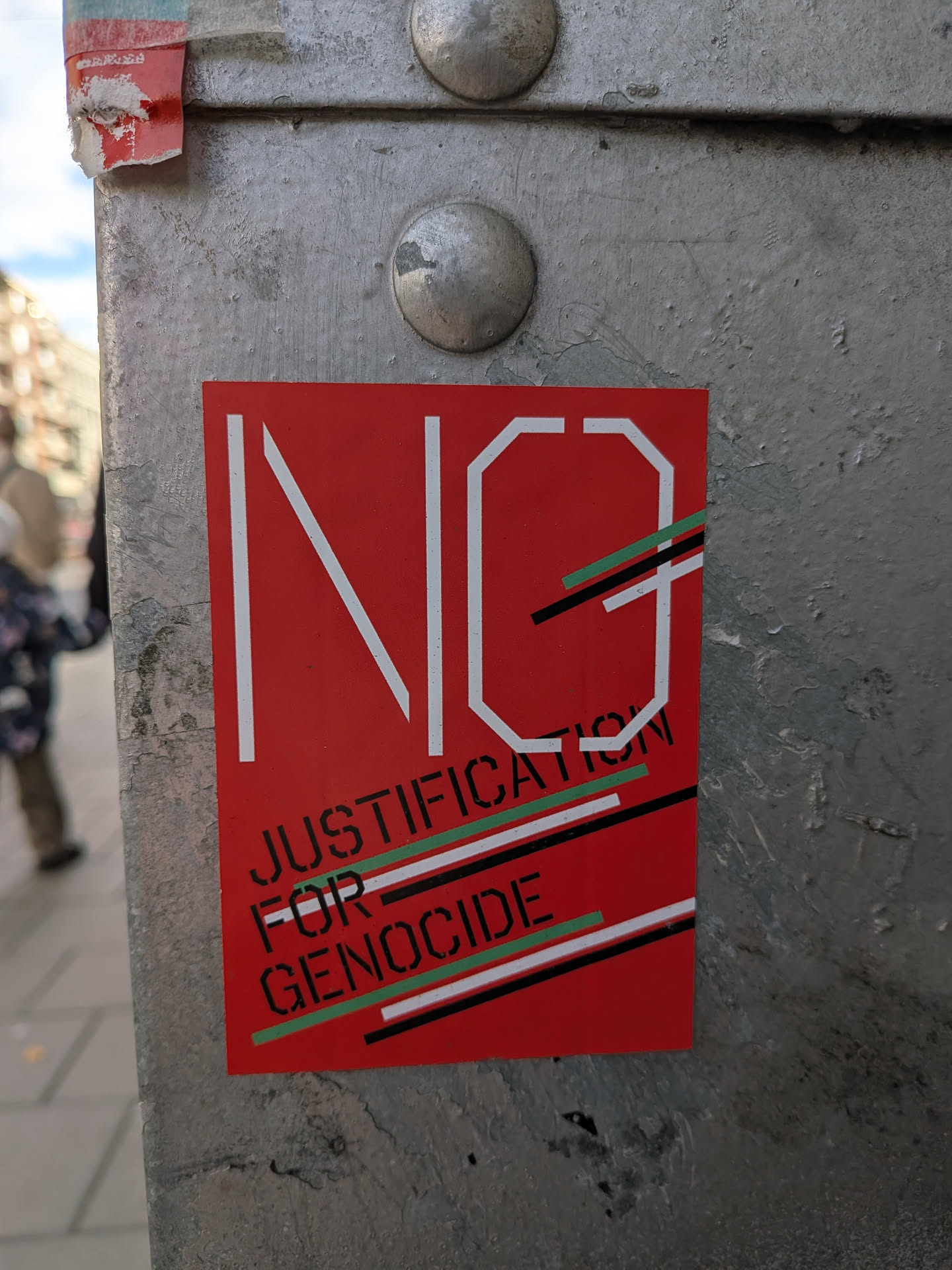 The sticker is predominantly red with white lettering. The word 'NO' is prominently displayed in large, stylized white block letters. Below 'NO', the text 'JUSTIFICATION FOR GENOCIDE' is written in smaller, black, stencil-like block letters. Diagonal lines in green, white, and black are layered across the lower portion of the sticker, adding a graphic element.