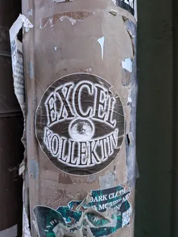 Street sticker A circular sticker featuring a prominent, stylized eye graphic at its center. Above the eye, the word "EXCEL" is written in a bold, distressed serif font with a white outline and dark fill. Below the eye, the word "KOLLEKTIV" is rendered in the same font and style. The background of the sticker is a dark, mottled grey or black, giving it a worn and textured appearance. The overall color scheme is monochrome, primarily black and white/off-white, with a grunge aesthetic. The sticker itself shows signs of wear, with slightly torn edges and creases, indicating it's affixed to a weathered surface.