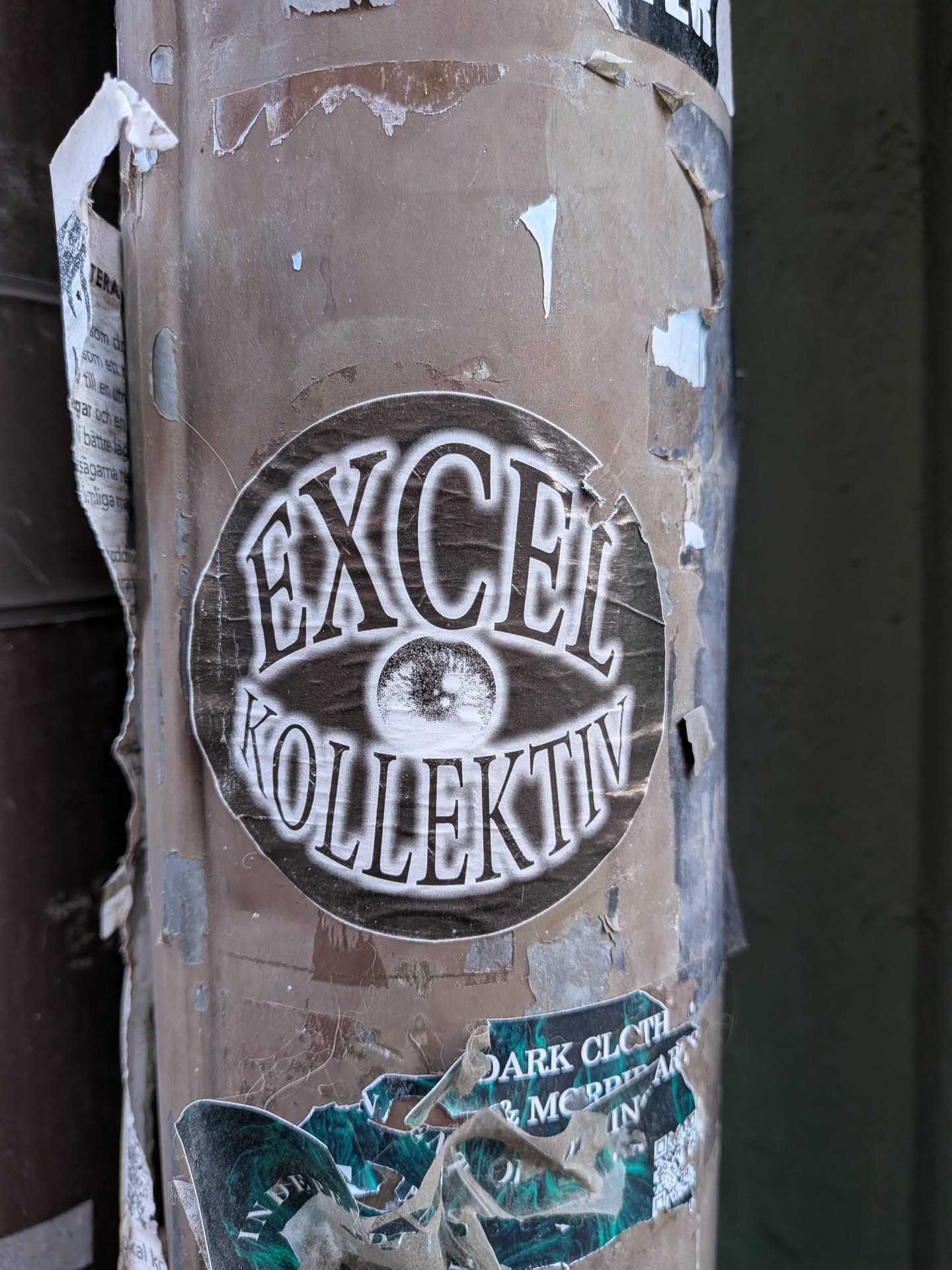A circular sticker featuring a prominent, stylized eye graphic at its center. Above the eye, the word "EXCEL" is written in a bold, distressed serif font with a white outline and dark fill. Below the eye, the word "KOLLEKTIV" is rendered in the same font and style. The background of the sticker is a dark, mottled grey or black, giving it a worn and textured appearance. The overall color scheme is monochrome, primarily black and white/off-white, with a grunge aesthetic. The sticker itself shows signs of wear, with slightly torn edges and creases, indicating it's affixed to a weathered surface.