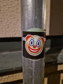 A round sticker featuring a cartoon clown emoji with big blue eyes and a red smile. The sticker is on a dark background with a light green circle behind it. Some text is partially visible on the edges of the sticker.